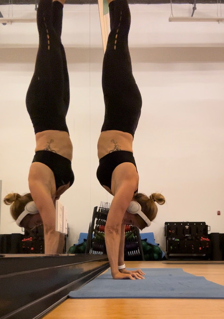 Shelley doing a handstand in front of a large mirror in a gym like studio while listening to music through headphones.